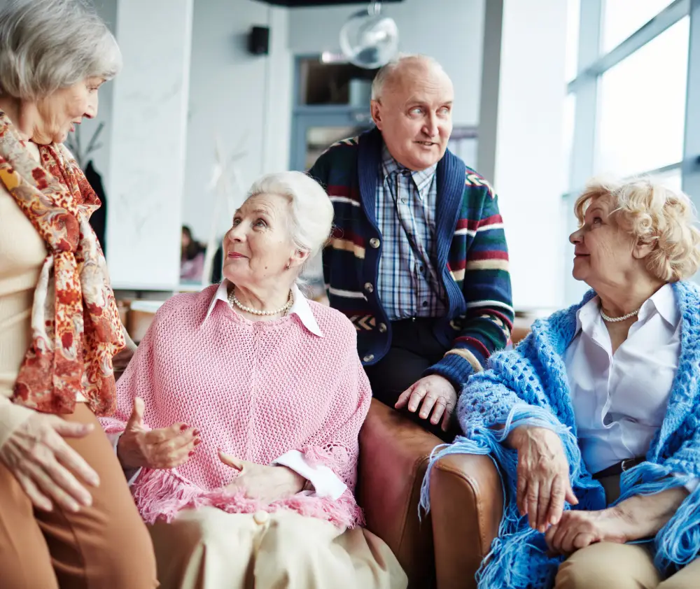 Aged-adults-having-conversation-indoors-1 A courtyard with a grassy area and a patio with a table and chairs, between two buildings with dark roofs and siding