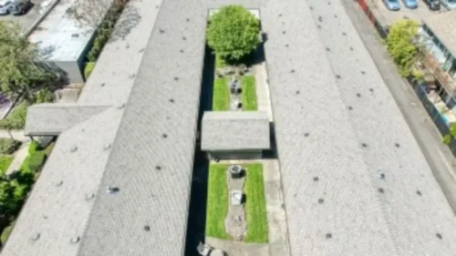 An aerial view of two rows of buildings with gray roofs, separated by a grassy courtyard with a tree and a fountain