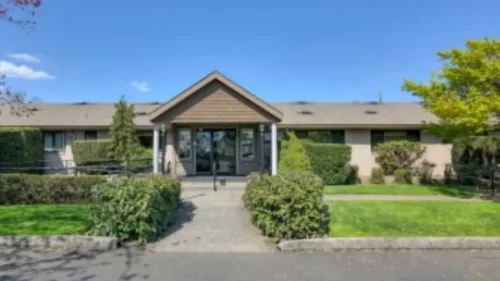 The front entrance of a building with a covered walkway, a stone facade, and a walkway surrounded by green bushes